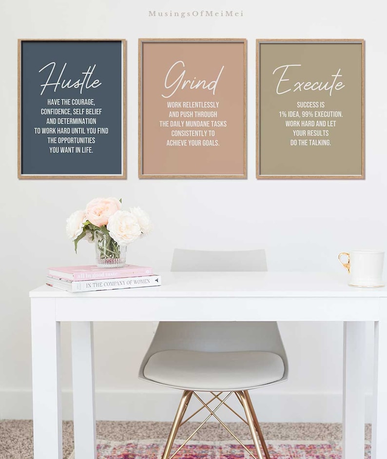 May include: Three motivational posters with a light brown wood frame. The posters have a light blue, light brown, and light gray background. The text on the posters reads "Hustle", "Grind", and "Execute". The posters are hanging on a white wall above a white desk with a light gray chair and a vase of white and pink flowers.