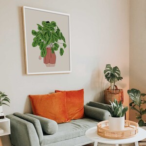 May include: A framed art print depicting a person with a plant head, wearing shorts. The artwork is in a white frame and hangs on a neutral wall. The room also includes a green sofa with orange cushions, and other plants.