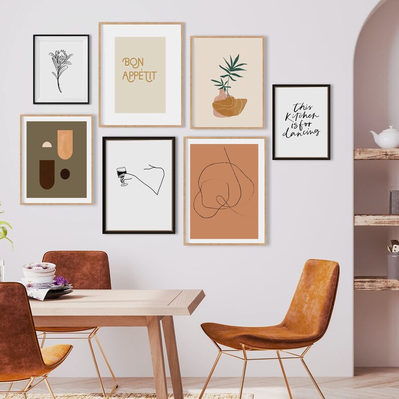 May include: A set of six framed prints with various designs, including abstract shapes, a plant, a glass of wine, and text. The prints are hung on a white wall above a wooden table with two brown chairs.