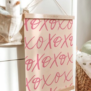 Puede incluir: Una impresi&oacute;n rosa y blanca con la palabra "XOXO" repetida en una fuente de pinceladas. La impresi&oacute;n est&aacute; enmarcada en un marco de madera natural y colgada en una pared blanca.