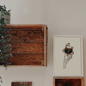 May include: A white framed print with a black and white illustration of a skeletal hand holding a bottle with a black orb and orange ribbon. The bottle has a small black hat on top.