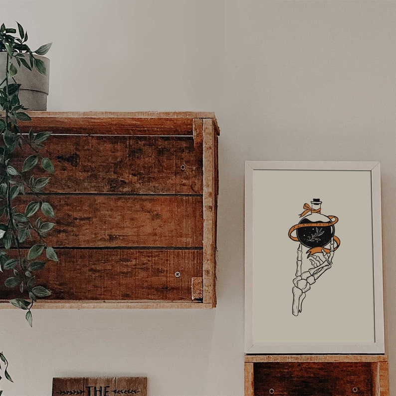 May include: A white framed print with a black and white illustration of a skeletal hand holding a bottle with a black orb and orange ribbon. The bottle has a small black hat on top.