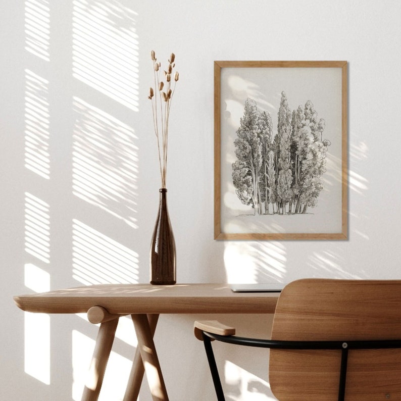 May include: A black and white line drawing of a group of trees in a wooden frame hangs on a white wall above a wooden desk with a chair. Dried flowers are in a brown glass vase on the desk.