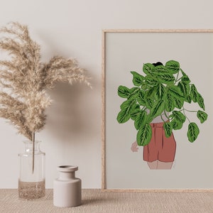 May include: A framed art print depicting a person with a large green plant obscuring their head and shoulders. The person wears a light grey top and rust-coloured shorts. The artwork is set against a neutral background.