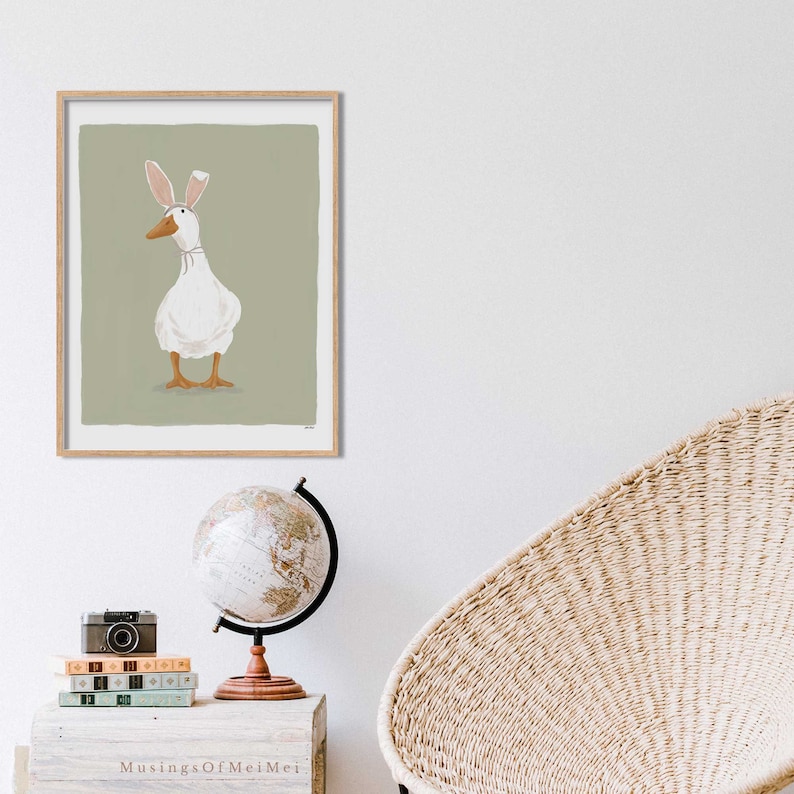 May include: A framed illustration of a white duck wearing bunny ears on a sage green background.