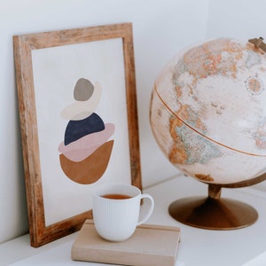 K&ouml;nnte beinhalten: Ein gerahmter abstrakter Kunstdruck mit Holzrahmen, der geschichtete Formen in Braun, Rosa, Marineblau und Grau zeigt. Eine wei&szlig;e Teetasse steht auf einem Tisch neben einem Globus und einem Notizbuch.