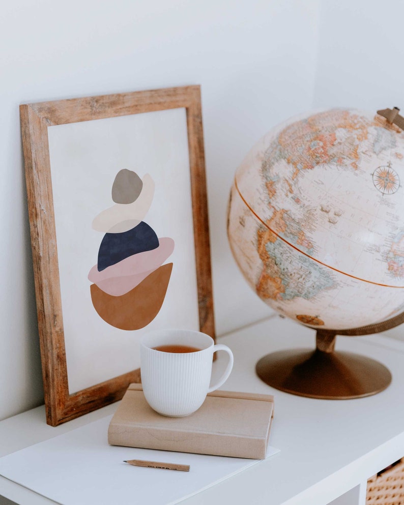 K&ouml;nnte beinhalten: Ein gerahmter abstrakter Kunstdruck mit Holzrahmen, der geschichtete Formen in Braun, Rosa, Marineblau und Grau zeigt. Eine wei&szlig;e Teetasse steht auf einem Tisch neben einem Globus und einem Notizbuch.