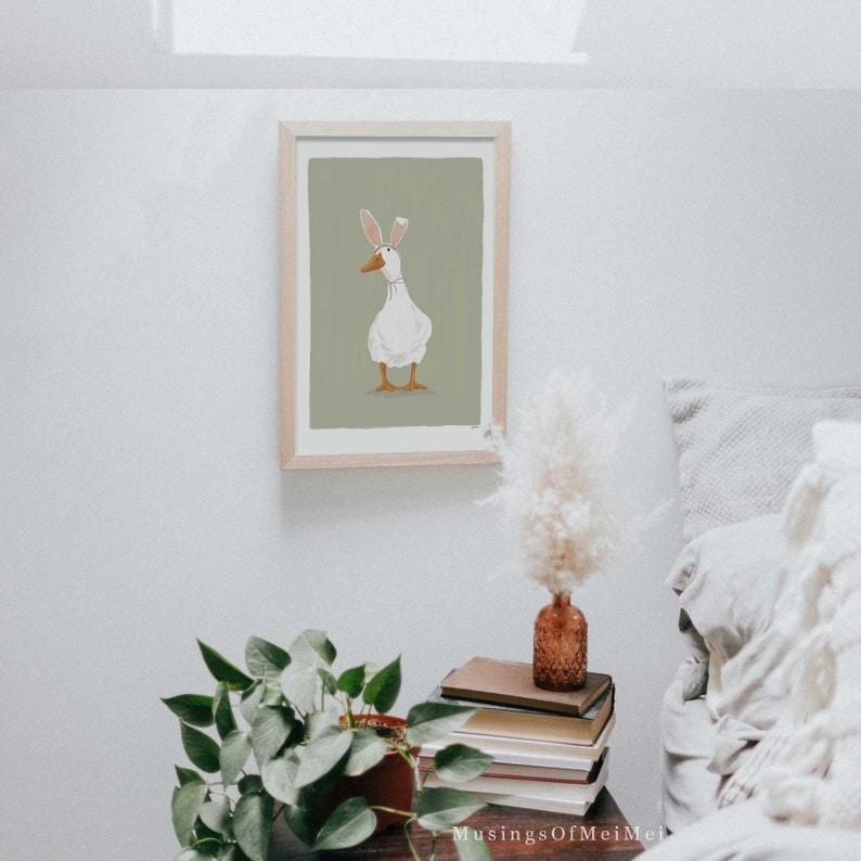 May include: A framed illustration of a white duck wearing bunny ears on a green background.
