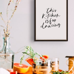 May include: A framed print with the text "This kitchen is for dancing" in black calligraphy on a white background. The frame is black with a gold trim. The print is hanging on a white wall.