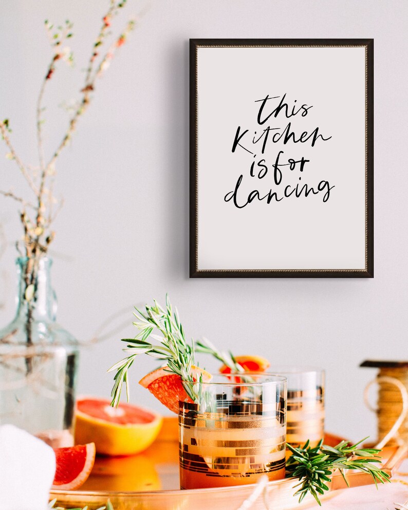 May include: A framed print with the text "This kitchen is for dancing" in black calligraphy on a white background. The frame is black with a gold trim. The print is hanging on a white wall.