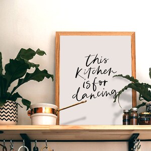May include: A framed print with the text "This kitchen is for dancing" in black calligraphy on a light gray background. The print is on a wooden shelf with plants and kitchen items.
