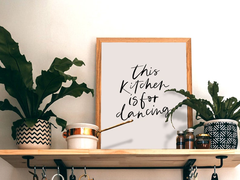 May include: A framed print with the text "This kitchen is for dancing" in black calligraphy on a light gray background. The print is on a wooden shelf with plants and kitchen items.
