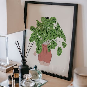 May include: A framed art print depicting a person with a plant obscuring their head. The person wears a white top and pink shorts. A black diffuser, perfume bottles, and a lamp are on a table.