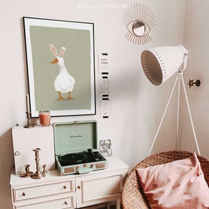 May include: A green framed print of a white duck wearing a pink bunny ear headband against a green background. The print is hanging on a white wall above a light green vintage record player. A white lamp with a tripod stand is in the background.