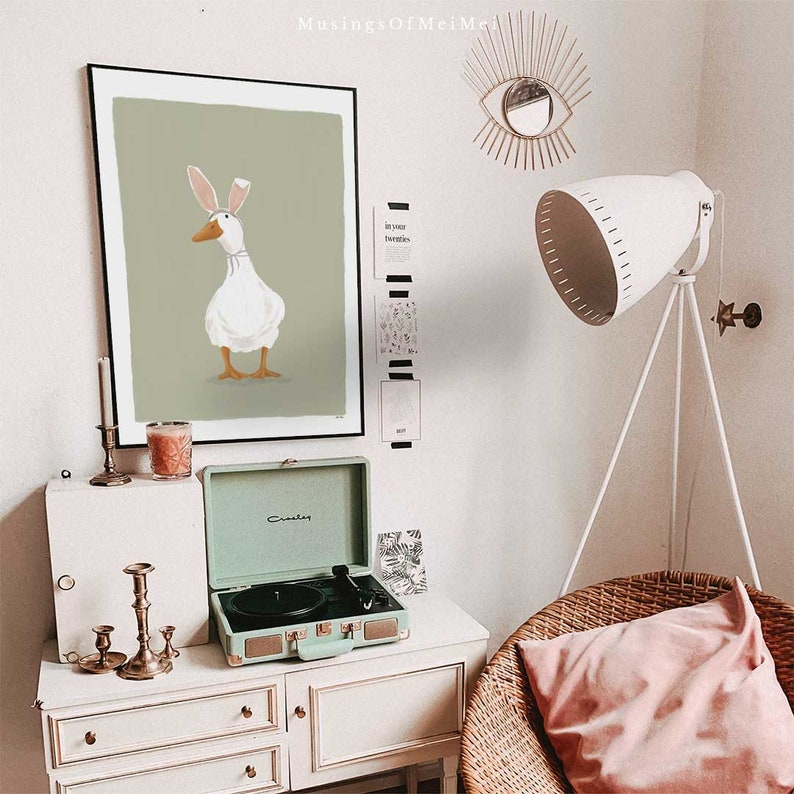 May include: A green framed print of a white duck wearing a pink bunny ear headband against a green background. The print is hanging on a white wall above a light green vintage record player. A white lamp with a tripod stand is in the background.