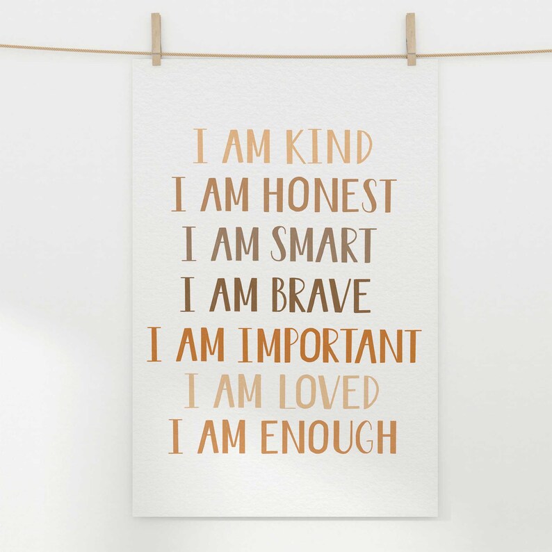 May include: A white poster with the words "I AM KIND, I AM HONEST, I AM SMART, I AM BRAVE, I AM IMPORTANT, I AM LOVED, I AM ENOUGH" printed in various shades of brown. The poster is hanging from a clothesline with a wooden clip.