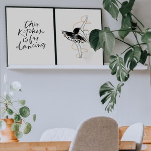 May include: Two framed prints on a white shelf. The print on the left says "This kitchen is for dancing". The print on the right features a black and white line drawing of a ballerina.