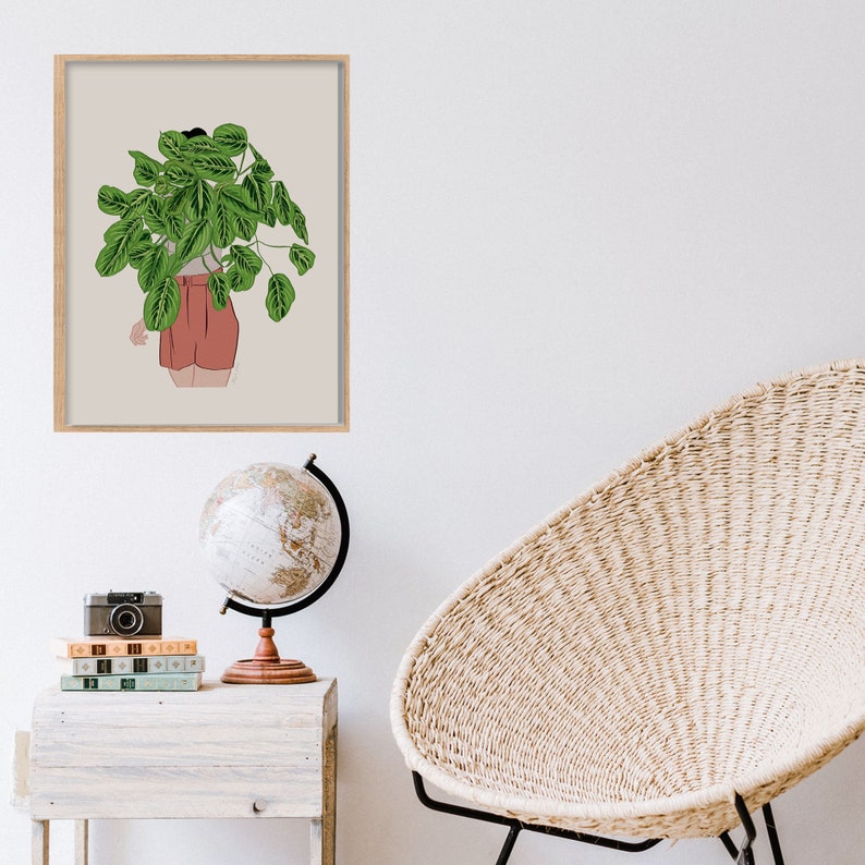 May include: A framed art print depicting a person with a large green plant obscuring their upper body, wearing red shorts. The artwork is in a light wood frame, set against a neutral background. A globe, books, and a camera are on a nearby table.