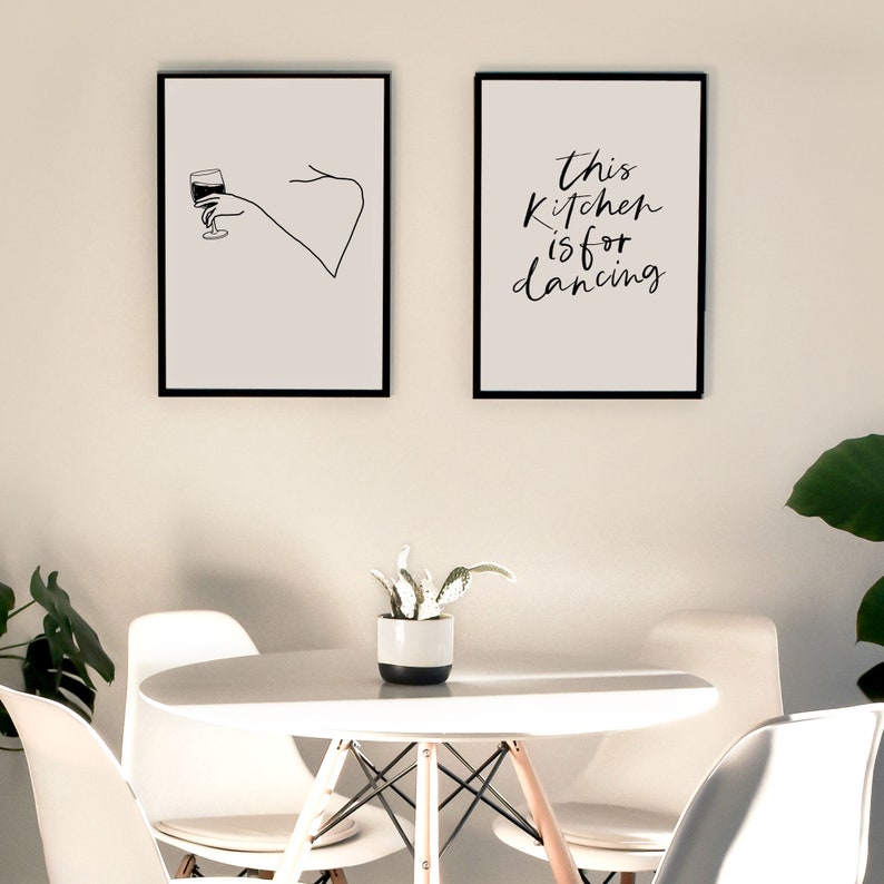 May include: Two black framed prints on a white wall. The print on the left shows a line drawing of a hand holding a glass of wine. The print on the right has the text "This kitchen is for dancing".
