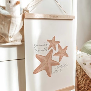 May include: A white framed print with a light brown background featuring three starfish and the text "Twinkle Twinkle Little Star". The print is hanging on a wall with a wooden dowel rod at the top and bottom.
