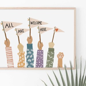 May include: A framed art print featuring hands holding pennant flags with the words "ALL ARE WELCOME HERE." The hands have various skin tones and patterned sleeves. One hand holds a paw print. The flags are beige with black text and a red heart.