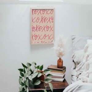 Puede incluir: Una impresi&oacute;n enmarcada con un fondo beige y un texto rosa "XOXO" repetido en un patr&oacute;n. La impresi&oacute;n est&aacute; colgada en una pared blanca sobre una mesita de noche de madera con una planta en maceta, libros y un jarr&oacute;n con flores secas.
