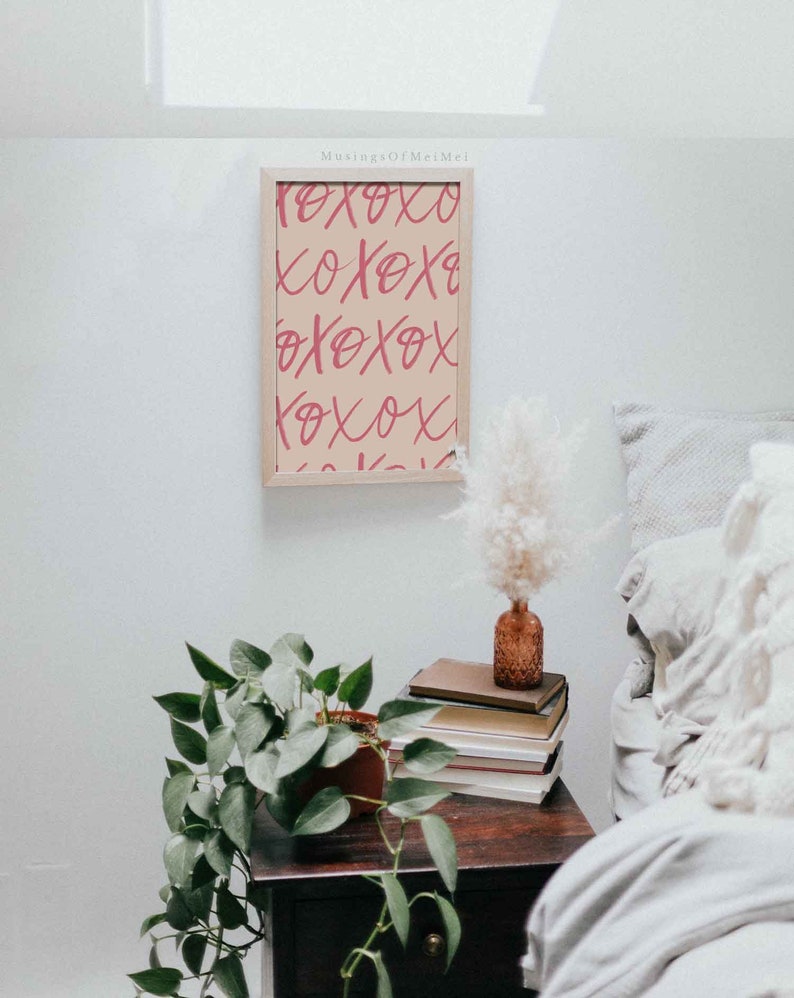 Puede incluir: Una impresi&oacute;n enmarcada con un fondo beige y un texto rosa "XOXO" repetido en un patr&oacute;n. La impresi&oacute;n est&aacute; colgada en una pared blanca sobre una mesita de noche de madera con una planta en maceta, libros y un jarr&oacute;n con flores secas.