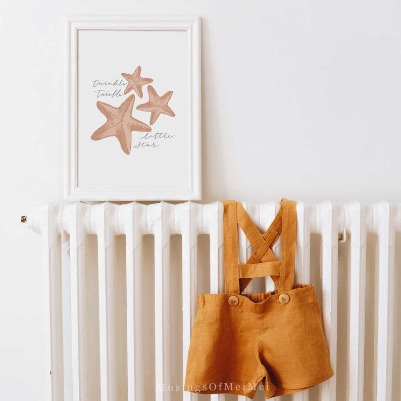 May include: A white framed print with a watercolor illustration of three starfish and the text "Twinkle Twinkle Little Star". The print is hanging on a white radiator with a pair of mustard yellow linen overalls hanging on the radiator.