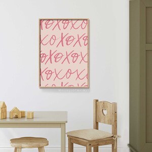 Puede incluir: Una impresi&oacute;n rosa y blanca con el texto "XOXO" repetido en un patr&oacute;n. La impresi&oacute;n est&aacute; enmarcada en un marco de madera clara y est&aacute; colgada en una pared blanca.
