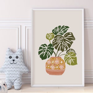 May include: A framed print of a green monstera plant in a terracotta pot with a geometric pattern. The background is a light beige color.