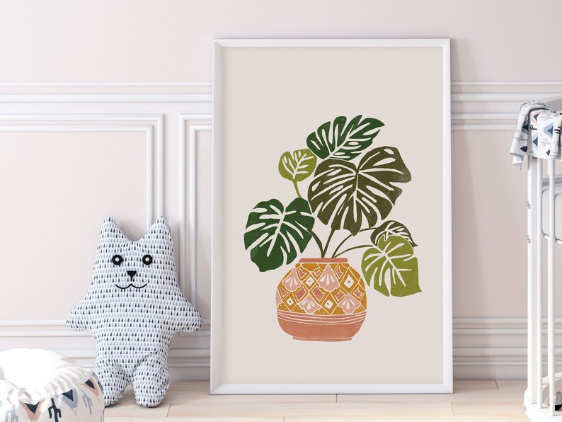 May include: A framed print of a green monstera plant in a terracotta pot with a geometric pattern. The background is a light beige color.