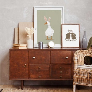 May include: Two framed prints on a wooden dresser. The print on the left shows a white duck wearing bunny ears. The print on the right shows two brown bunnies kissing with the text "Bunny Kisses Easter Wishes".
