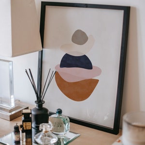 K&ouml;nnte beinhalten: Abstrakter Kunstdruck in einem schwarzen Rahmen mit geschichteten Formen in Braun-, Rosa-, Marine- und Graut&ouml;nen. Ein schwarzer Diffusor, Parf&uuml;mflaschen und eine Lampe stehen auf einem Tisch.