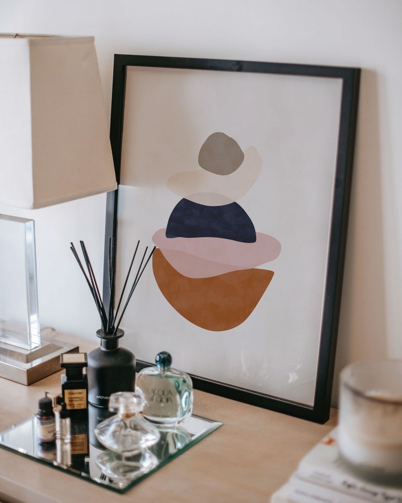 K&ouml;nnte beinhalten: Abstrakter Kunstdruck in einem schwarzen Rahmen mit geschichteten Formen in Braun-, Rosa-, Marine- und Graut&ouml;nen. Ein schwarzer Diffusor, Parf&uuml;mflaschen und eine Lampe stehen auf einem Tisch.