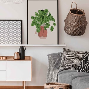 May include: A framed art print depicting a person with a large green plant obscuring their head and shoulders. The person is wearing a white top and coral shorts. The artwork is displayed on a white shelf with other decorative items.