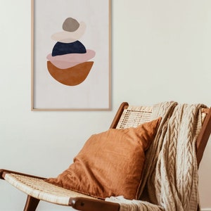 K&ouml;nnte beinhalten: Abstrakter Kunstdruck in einem Holzrahmen. Das Kunstwerk zeigt geschichtete Formen in Braun-, Marine-, Rosa- und Beiget&ouml;nen. Der Druck h&auml;ngt &uuml;ber einem Holzstuhl mit gewebtem Sitz, einem orangefarbenen Kissen und einer beigefarbenen Strickdecke.