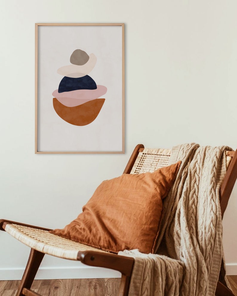 K&ouml;nnte beinhalten: Abstrakter Kunstdruck in einem Holzrahmen. Das Kunstwerk zeigt geschichtete Formen in Braun-, Marine-, Rosa- und Beiget&ouml;nen. Der Druck h&auml;ngt &uuml;ber einem Holzstuhl mit gewebtem Sitz, einem orangefarbenen Kissen und einer beigefarbenen Strickdecke.