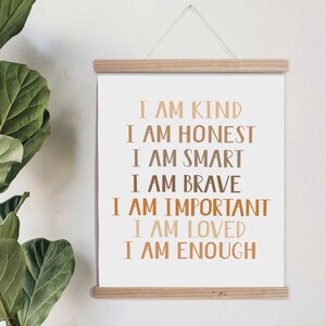 May include: A motivational art print with the words "I AM KIND, HONEST, SMART, BRAVE, IMPORTANT, LOVED, ENOUGH" in brown and tan lettering. The print is displayed on a white background and held by a wooden frame, with a hanging string.