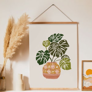 May include: A framed print of a green and brown plant in a pink and yellow patterned pot. The print is hanging on a white wall with a vase of dried pampas grass in front of it.