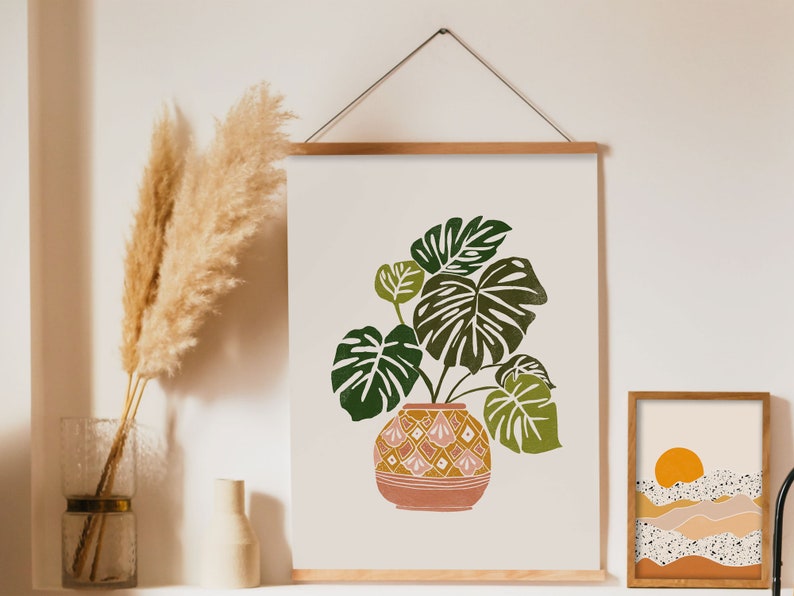 May include: A framed print of a green and brown plant in a pink and yellow patterned pot. The print is hanging on a white wall with a vase of dried pampas grass in front of it.