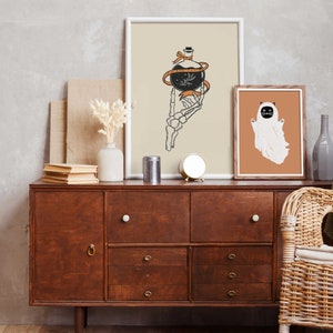 May include: Two framed prints on a wooden dresser. The print on the left features a skeletal hand holding a potion bottle with a ribbon tied around it. The print on the right features a white ghost with a black cat face.