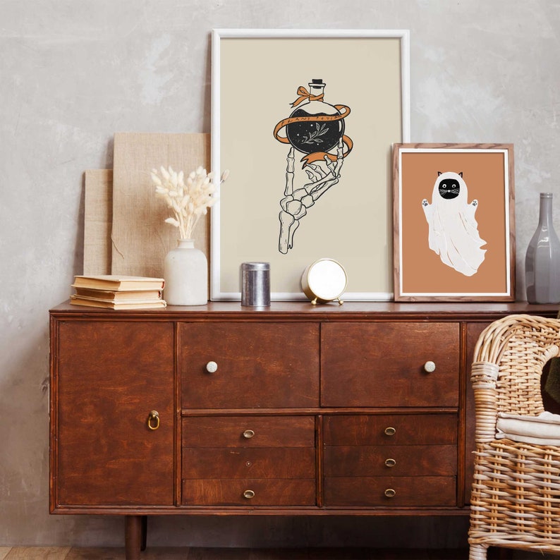 May include: Two framed prints on a wooden dresser. The print on the left features a skeletal hand holding a potion bottle with a ribbon tied around it. The print on the right features a white ghost with a black cat face.