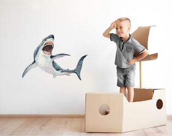 以下が含まれることがあります： グレーのシャツと青いショートパンツを着た子供が、船の形をした段ボール箱の中に立って、口を開けた大きなサメの壁のステッカーを見上げています。