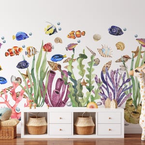 May include: A wall decal set featuring watercolor illustrations of various tropical fish, seashells, starfish, and aquatic plants. The scene includes a plush whale and a plush giraffe standing on a white storage unit with two woven baskets.