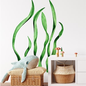 May include: A light blue whale plush toy is lying on a tan pillow in a wicker basket. The whale has large, blue eyes. The background features a white wall with a green watercolor seaweed design. There are small, colorful mushroom figurines on a white shelf.