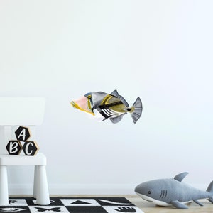 May include: A watercolor illustration of a grey, yellow, and blue triggerfish with black stripes. The fish is swimming to the right. The illustration is on a white wall with a white shelf and wooden blocks that spell "ABC".