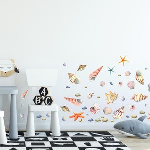 May include: A white wall with a variety of colorful seashells and starfish decals. The decals are arranged in a random pattern and are all different sizes. There are also some small, gray rocks scattered around the decals. The wall is in a room with a white chair and a white table.