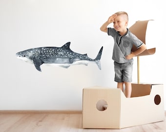 以下が含まれることがあります： 白壁に、グレーとブルーの色合いで描かれたジンベイザメの水彩イラスト。グレーのシャツとショートパンツを着た子供が、段ボールで作られた船の中に立ち、片手を目の上に置いてサメの方を見ています。