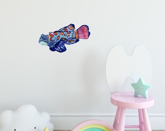 Op de afbeelding: Een kleurrijke vis-muursticker met blauwe, oranje en rode accenten. De sticker is geplaatst op een witte muur met een roze kruk en regenboogkleurige houten blokken op de voorgrond.