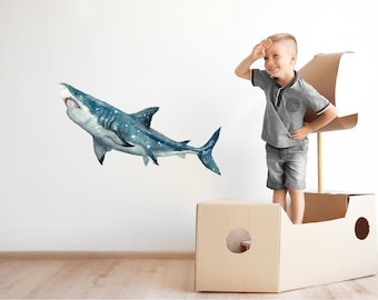 以下が含まれることがあります： 若い男の子が船の形をした段ボール箱の中に立って、青と白のシャークの壁のステッカーを見上げています。サメは星形の模様をしていて、男の子に向かって泳いでいます。